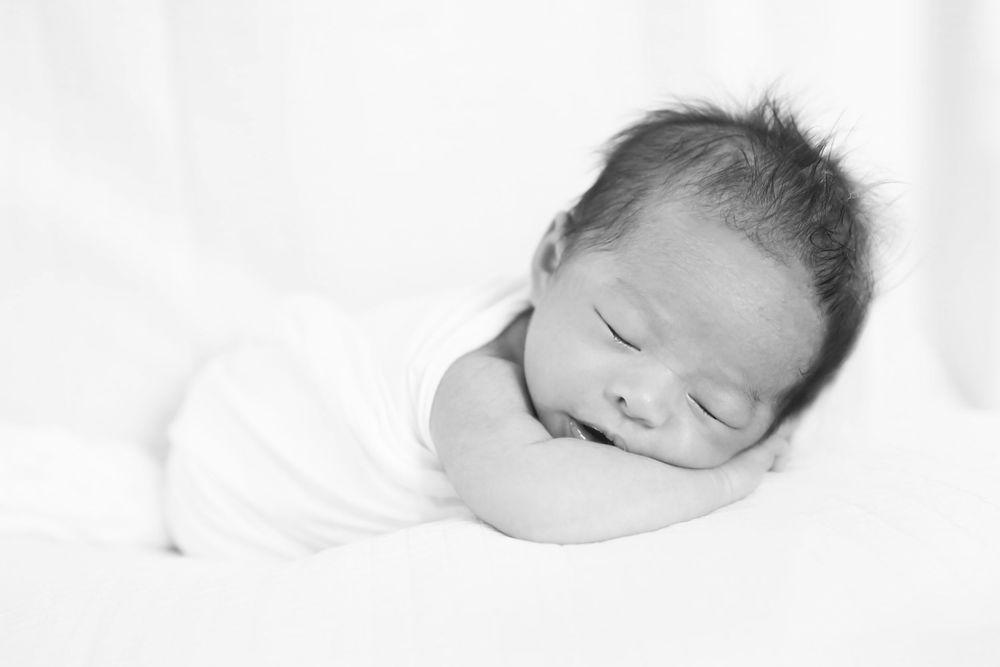 A peaceful sleeping newborn baby wrapped in white cloth rests on soft bedding in a gentle black and white portrait.