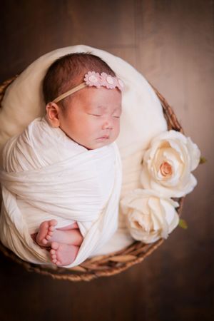Sleeping newborn wrapped in white cloth rests peacefully in round woven basket with soft peach rose decoration.
