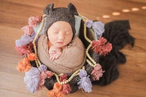 Sleeping newborn wrapped in soft brown swaddle with knit gray hat lying among colorful pom pom decorations on wooden floor.