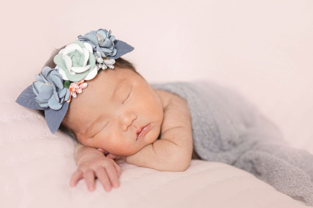 Peaceful sleeping newborn wearing delicate blue and white floral headband wrapped in soft gray blanket.