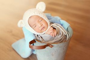 Sleeping baby wearing white bear hat nestles peacefully in vintage metal bucket on hardwood floor.