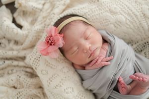 A sleeping newborn wrapped in soft gray fabric on a cream knitted blanket, adorned with a pink flower headband.