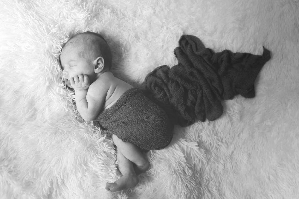 A sleeping baby lies on soft white fur wearing dark colored onesie in black and white photograph.