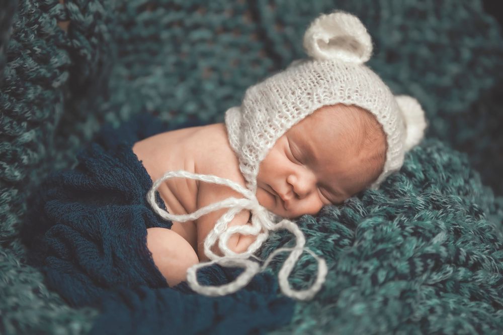 Newborn baby sleeping peacefully in a knit white bonnet amidst a dark blue fabric backdrop.