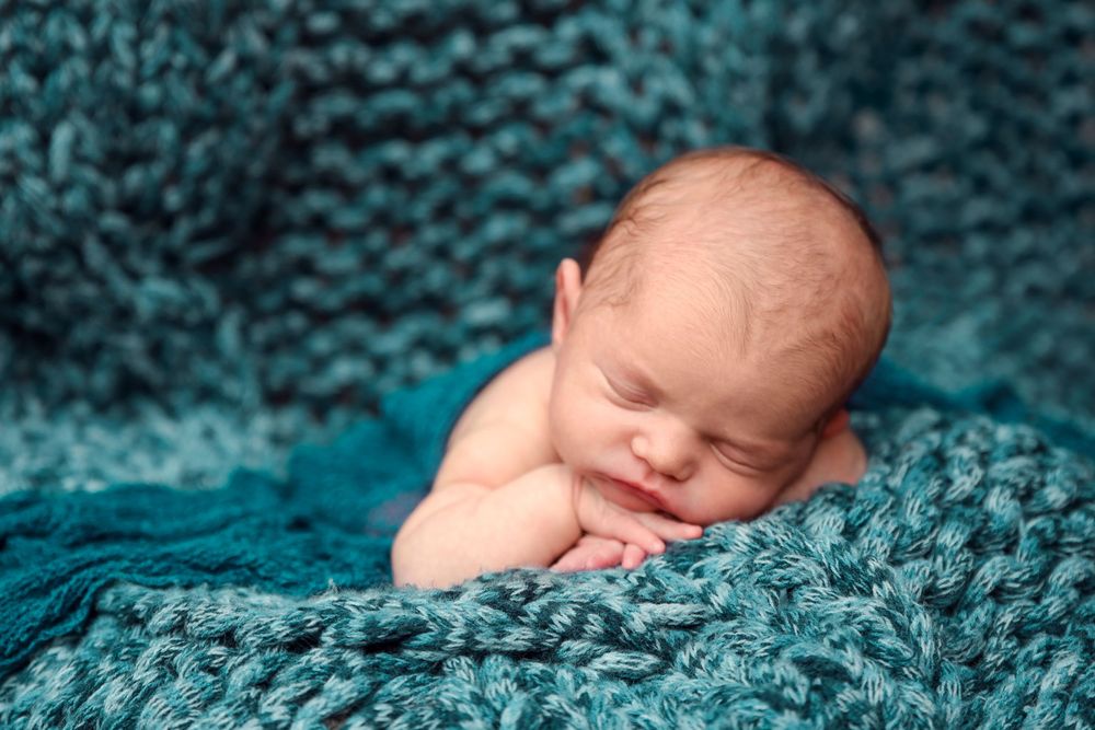 Peaceful sleeping newborn wrapped in cozy turquoise knit blanket during professional portrait session.