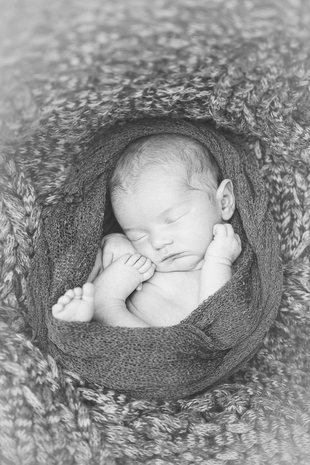 Newborn baby sleeping peacefully wrapped in soft gray knit blanket in black and white photograph.