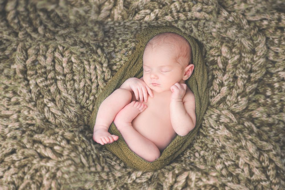 Newborn baby sleeps peacefully curled up on soft textured beige knit blanket during professional photography session.