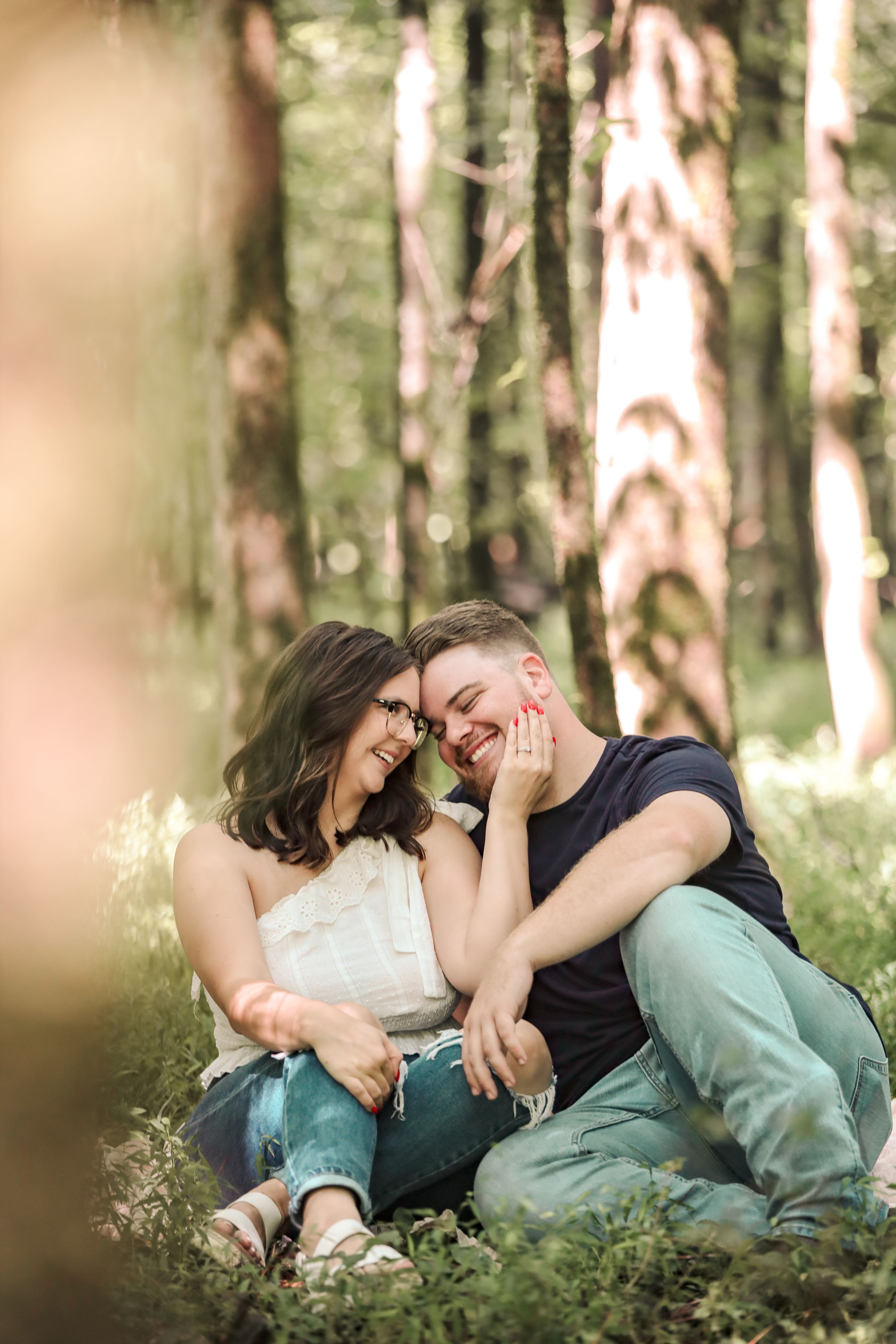 A couple sits together laughing and cuddling on grass in a sunlit forest with tall trees in the background.