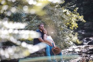 A couple shares an intimate moment by a stream in a dreamy forest setting with soft bokeh lighting effects.