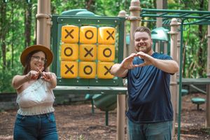 Two people make heart shapes with their hands while standing in front of a yellow tic-tac-toe game board at a playground.
