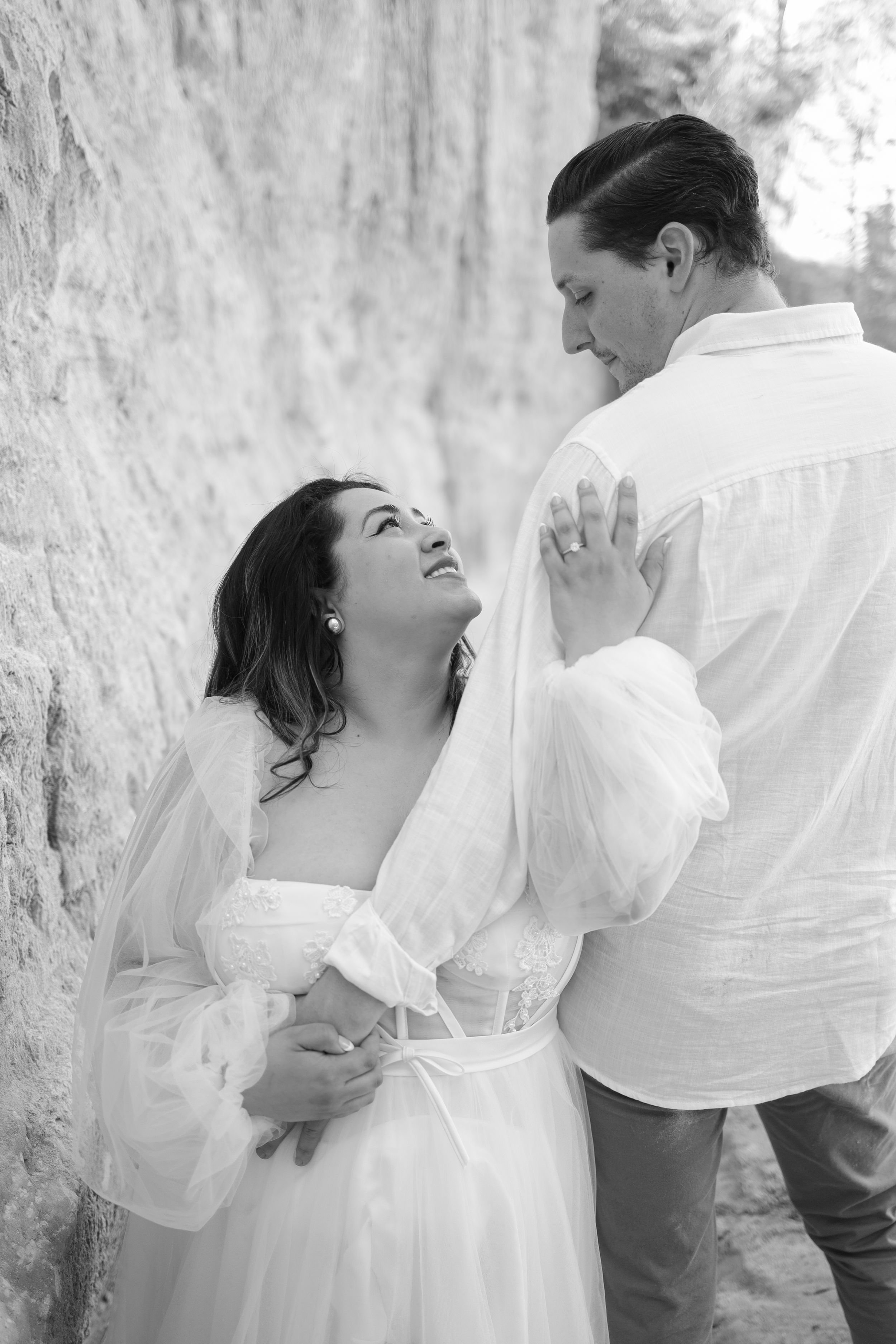 Black and white romantic engagement photo series showing a couple in white clothing posing playfully against a stone wall.