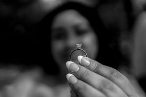 A close-up black and white photo shows hands holding up a delicate engagement ring against a blurred background.