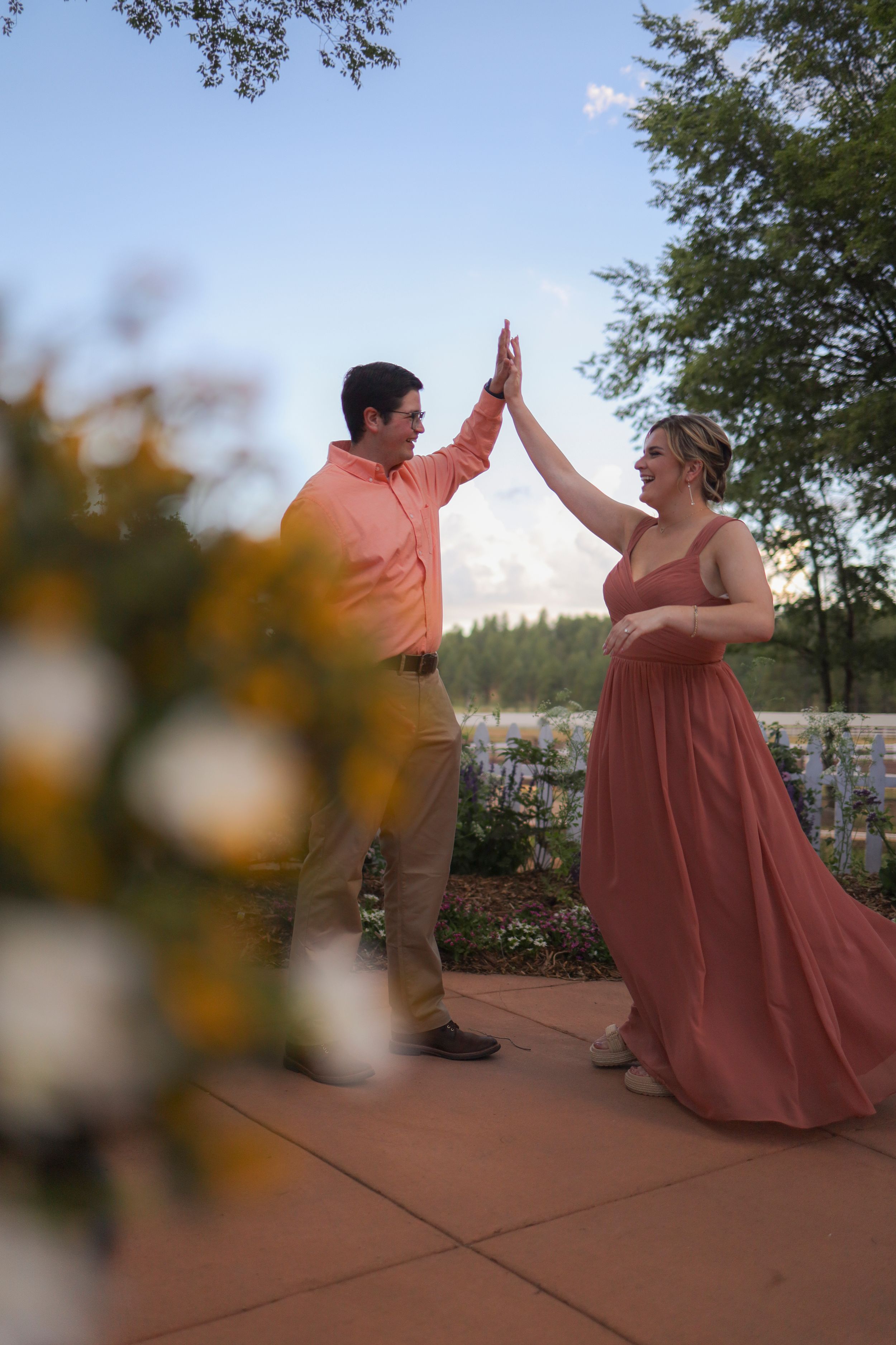 Joyful couple in wedding attire sharing a high five on an outdoor patio with yellow flowers in foreground.