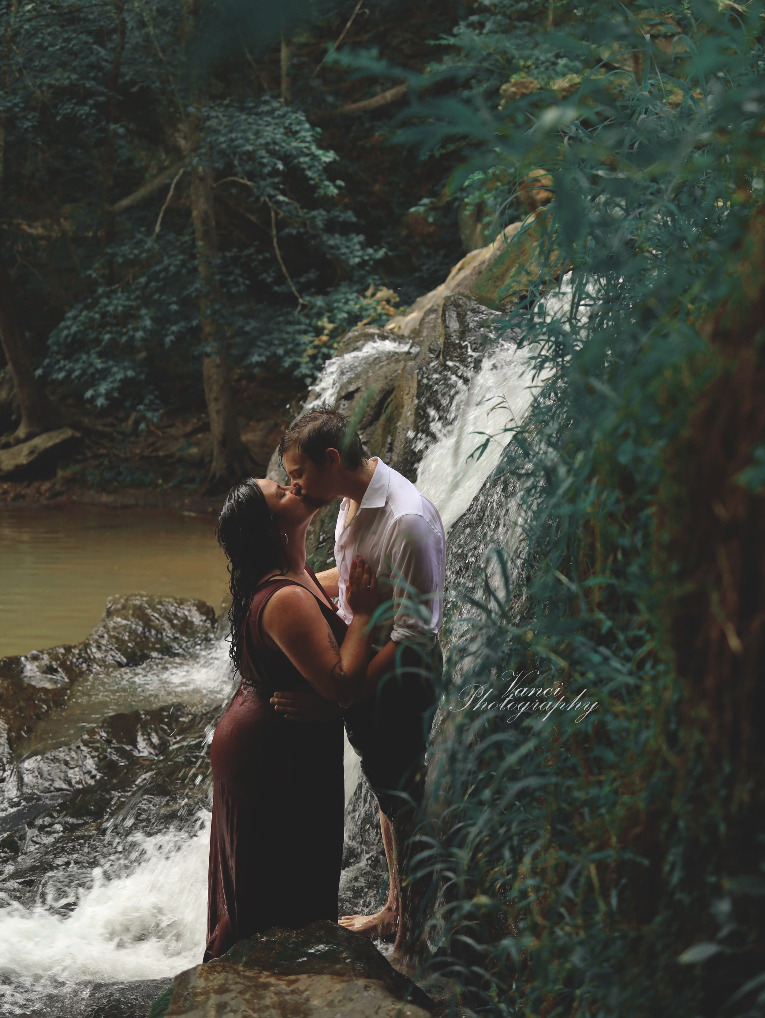 A romantic couple embraces by a cascading waterfall in a lush forest setting during a dreamy photoshoot.