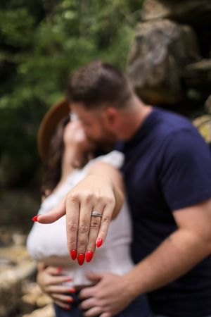 Close up of an engagement ring on a finger with bright red nail polish as a couple embraces in the background.