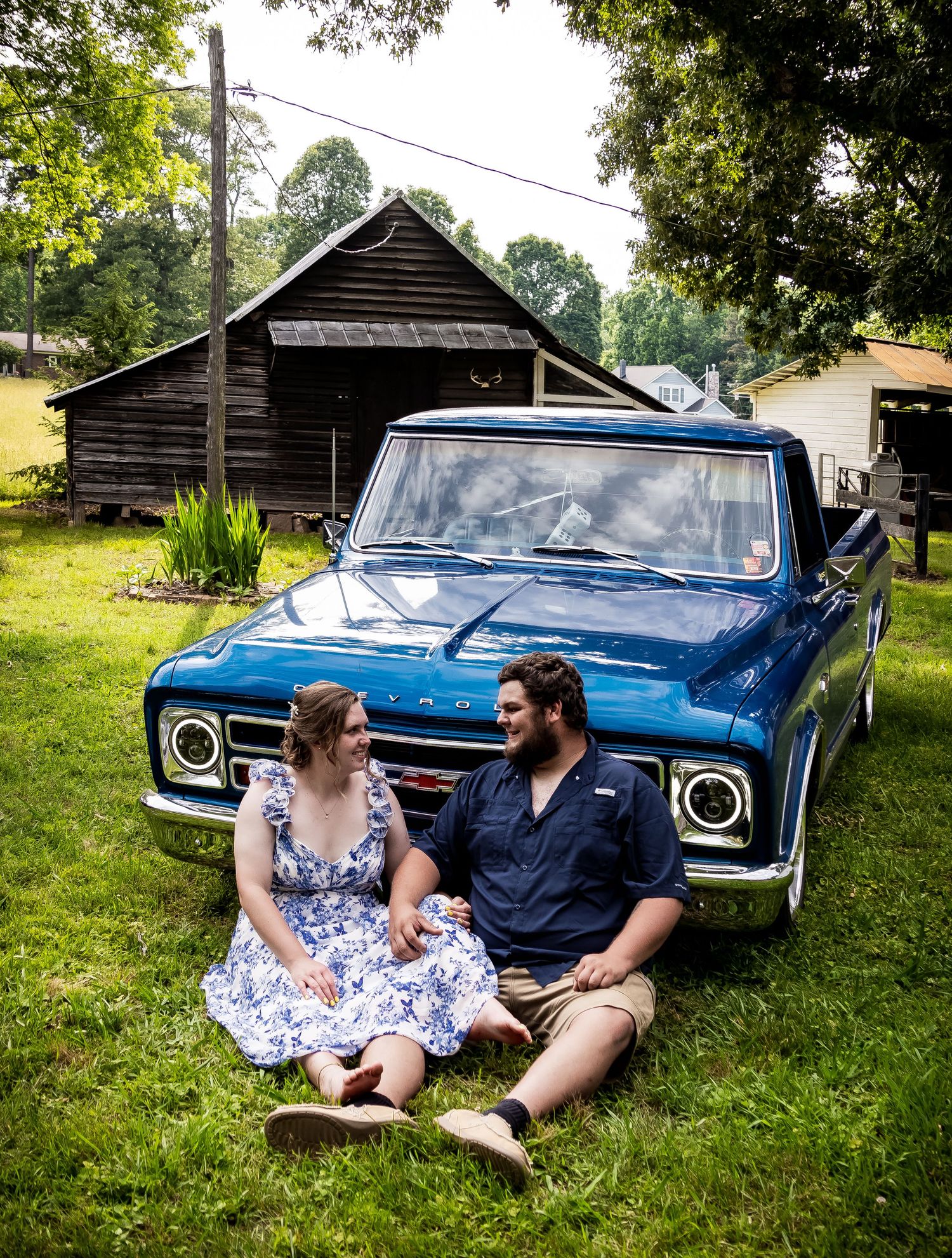 Classic blue pickup truck parked in front of rustic wooden cabin with occupants sitting by the front grille in a rural setting.