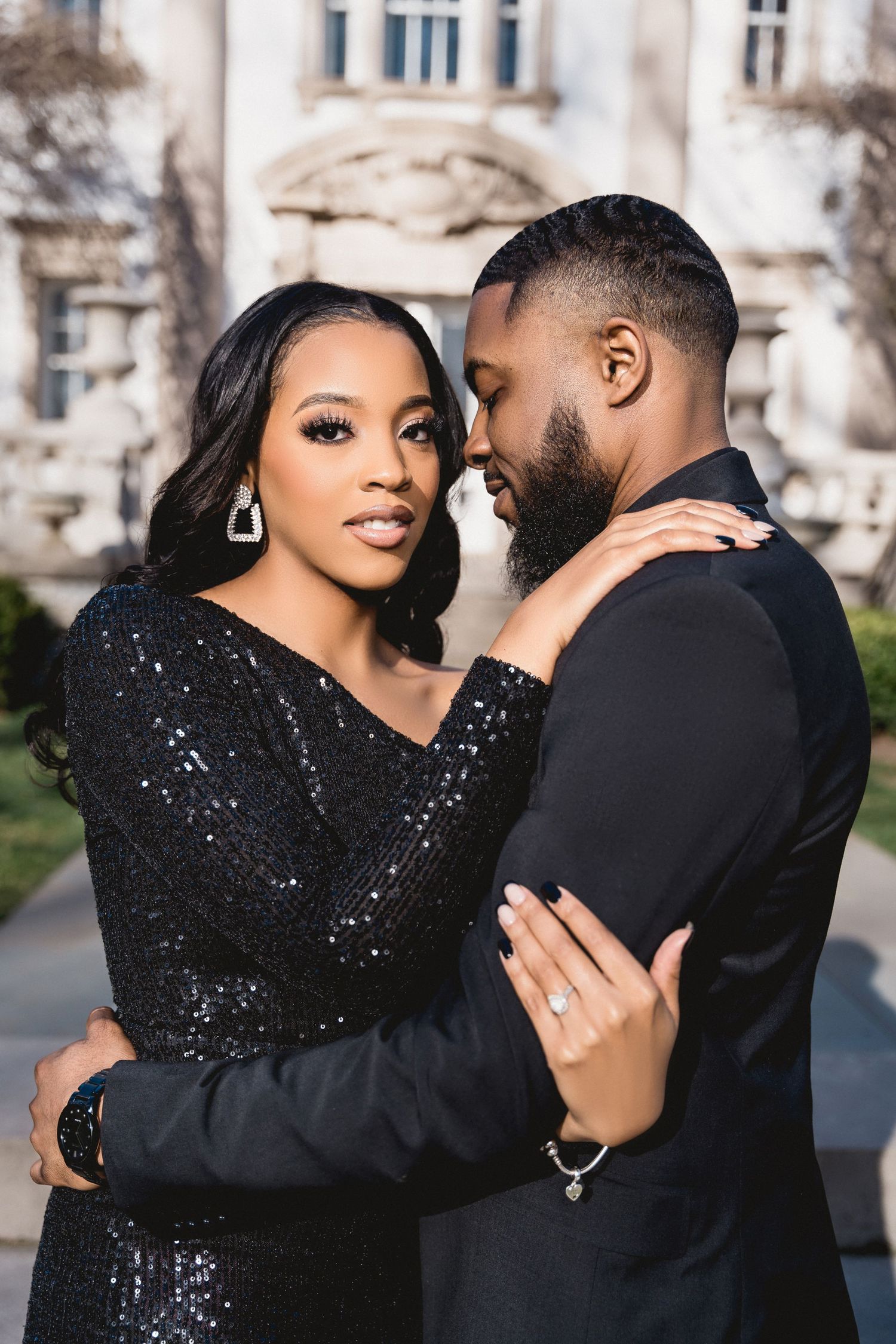 Elegant couple in black formal attire embrace outdoors against ornate architecture during romantic photo session.