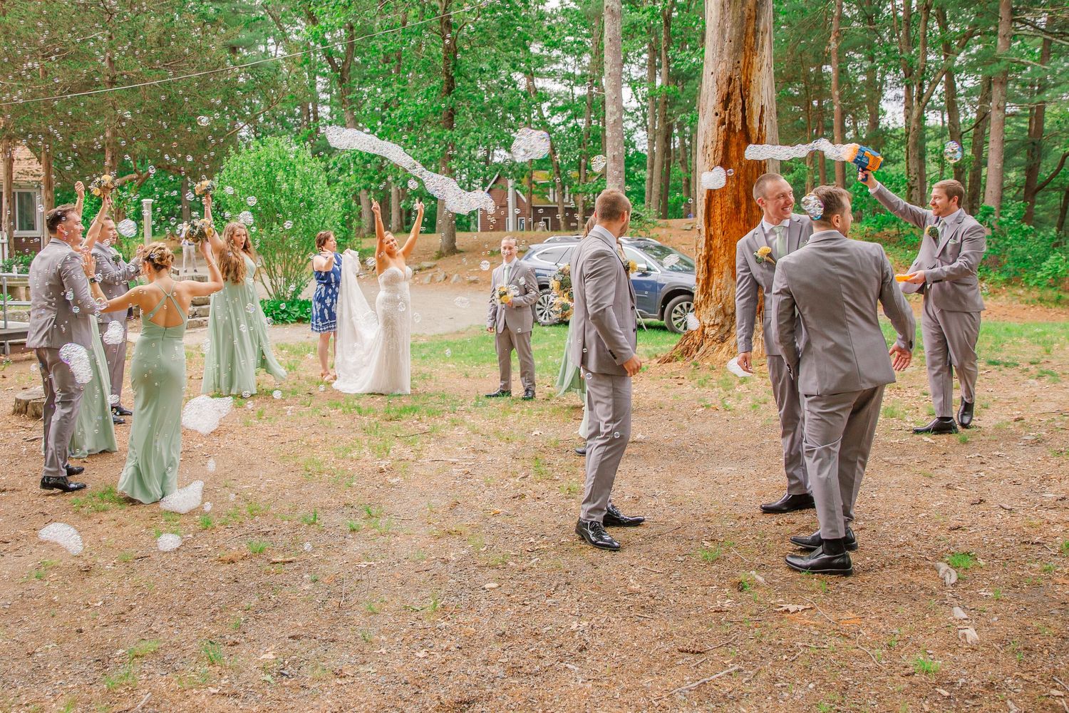 Wedding party celebrates outdoors in a wooded forest clearing with people in suits and dresses gathered together.