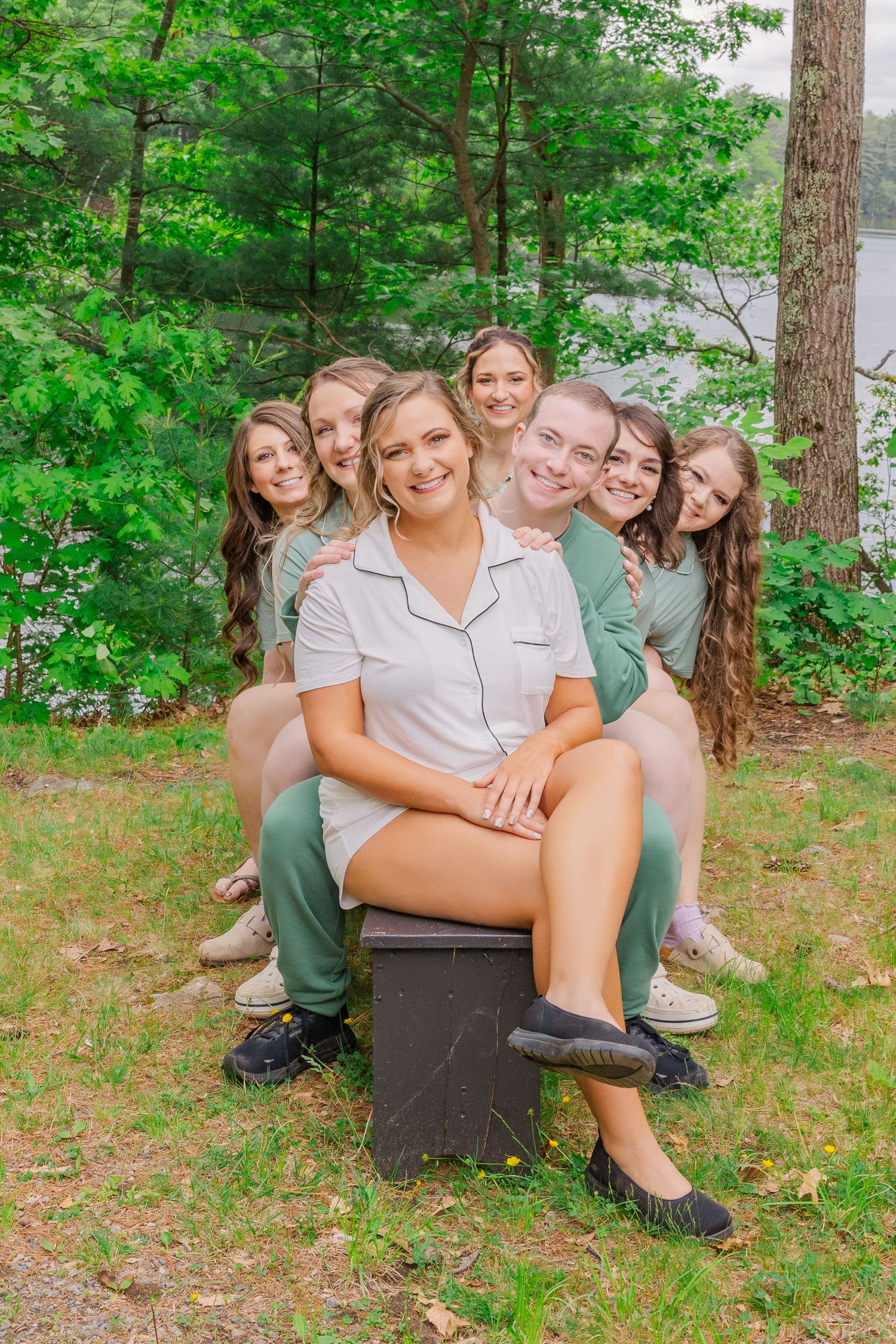 Group sitting together on a bench outdoors in a wooded area wearing casual summer clothing.