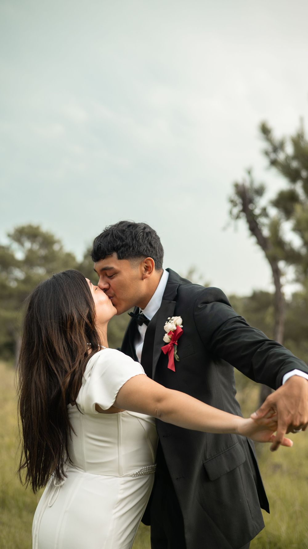 A couple shares a romantic kiss in formal wedding attire while standing in a natural outdoor field setting.