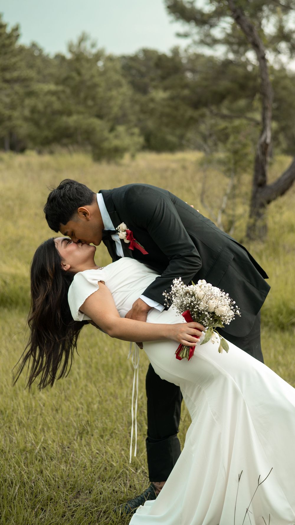 A couple shares a romantic dip kiss in a grassy field, the bride in white holding a bouquet and groom in black suit.