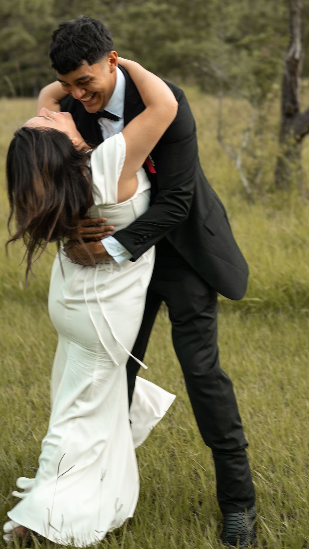 A couple in formal wedding attire shares an intimate dance together in a sunlit meadow with tall grass.