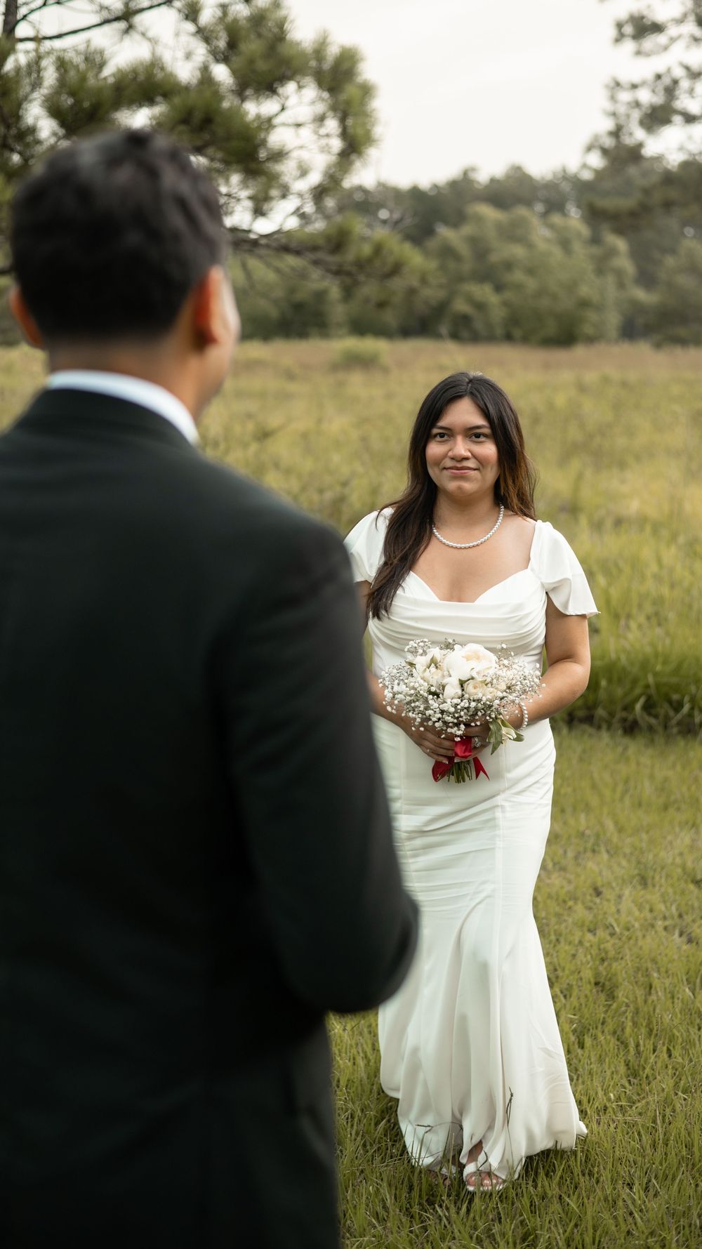 Bride in a white mermaid wedding dress holds bouquet while standing in a grassy field during portrait session.