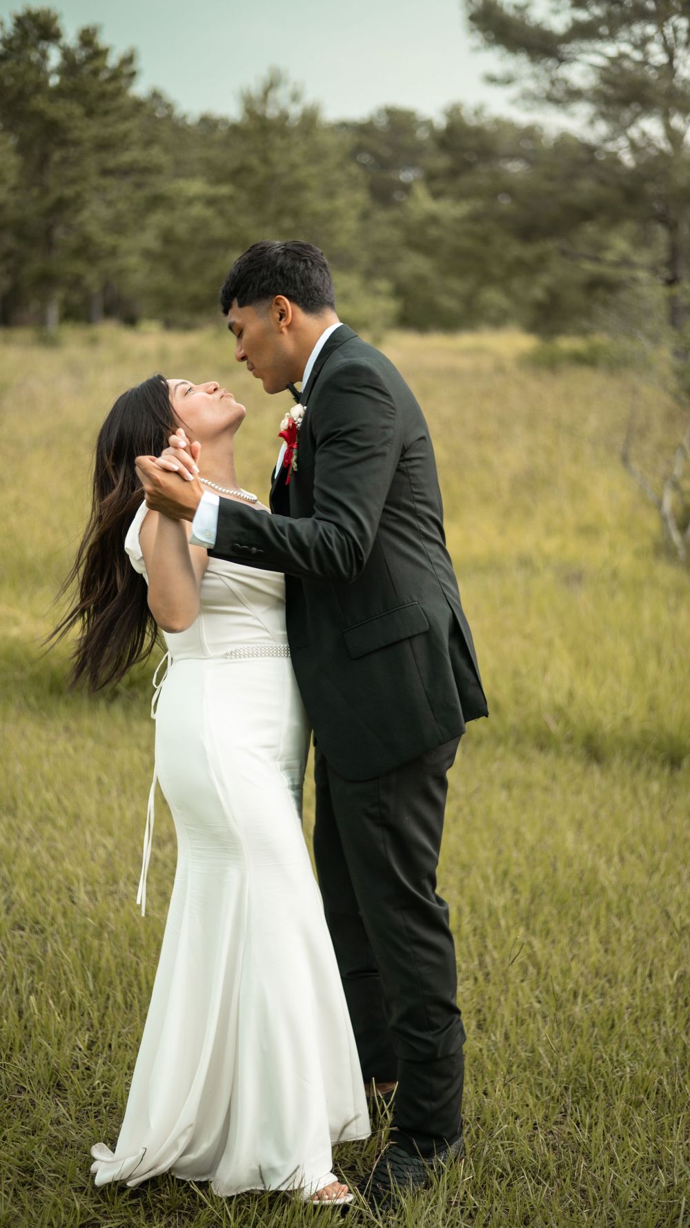 Newlywed couple shares romantic moment in open field with pine trees in background.