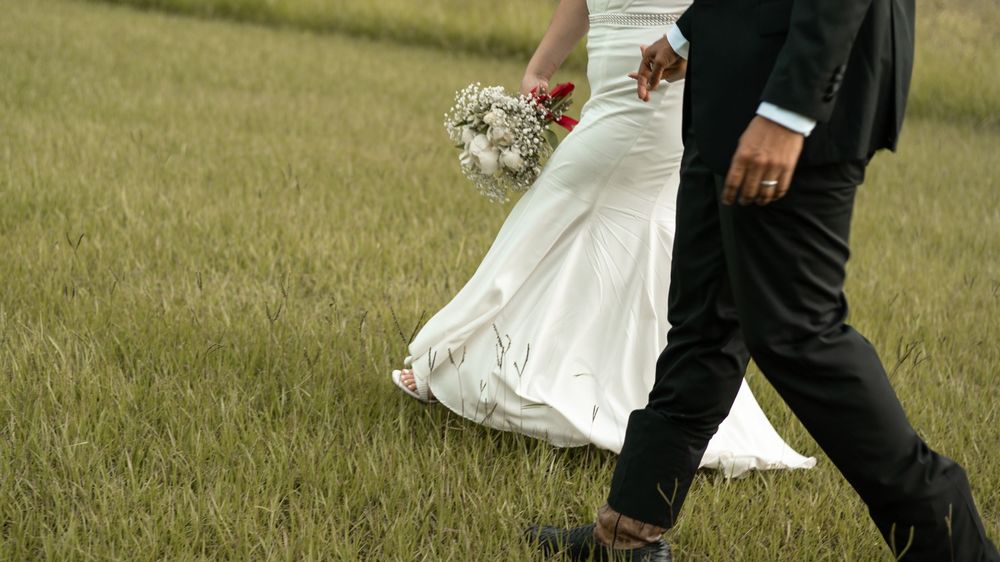 A couple walking through a grassy meadow with the dress and bouquet in white against a dark suit and green background.
