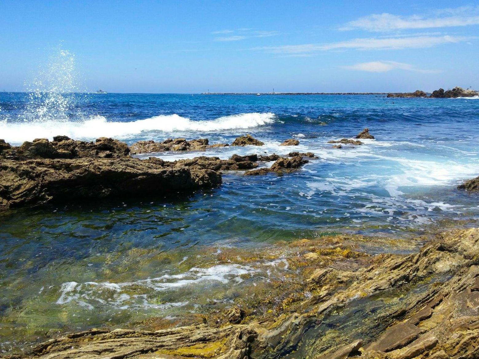 Ocean waves crash against rocky coastal formations on a sunny day with clear blue skies and turquoise waters.