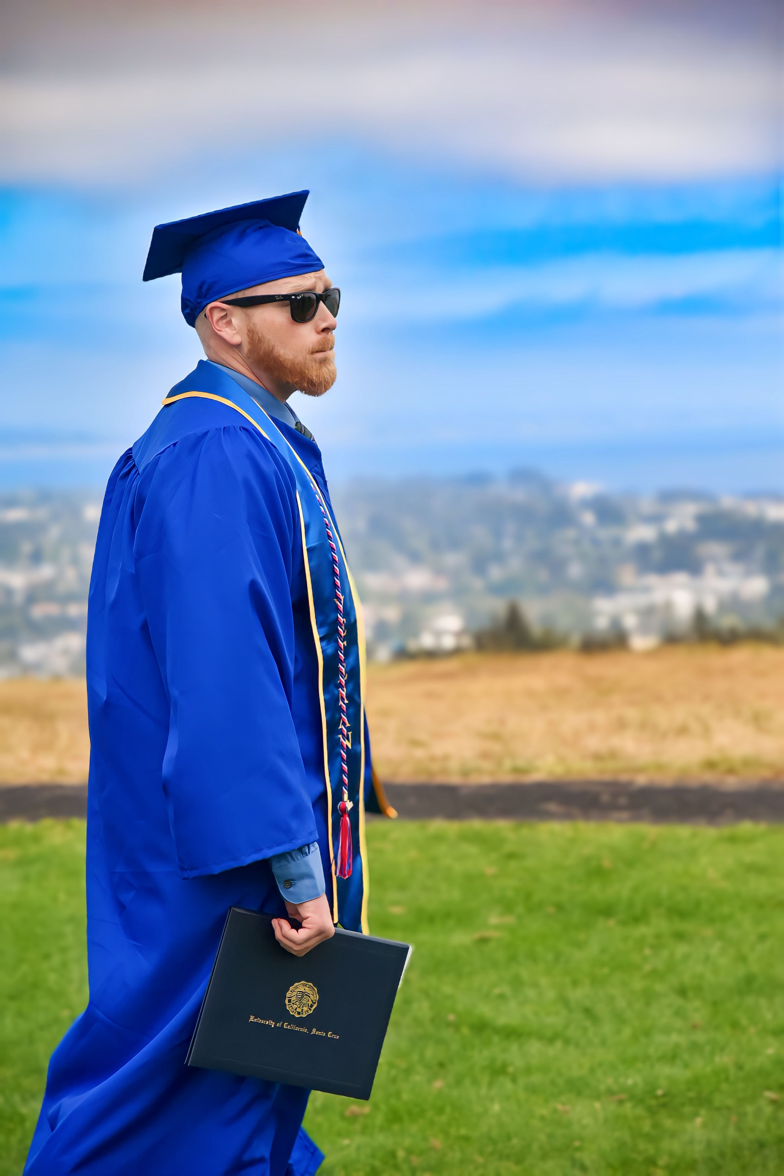 Graduate in blue cap and gown holds diploma while standing on hilltop overlooking city landscape on sunny day.