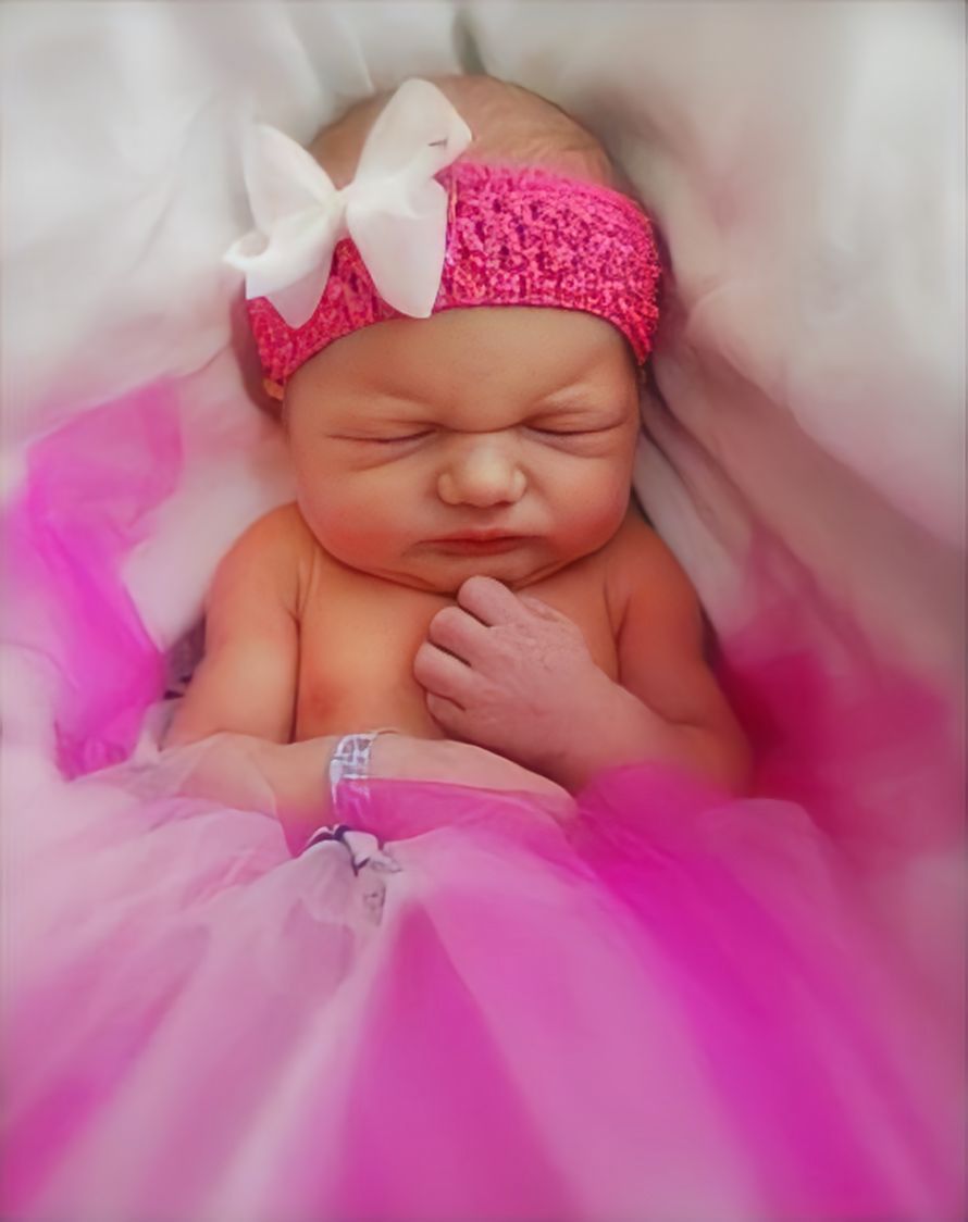 Sleeping newborn wearing pink tutu and crochet headband with white bow rests on soft pink and white fabric.
