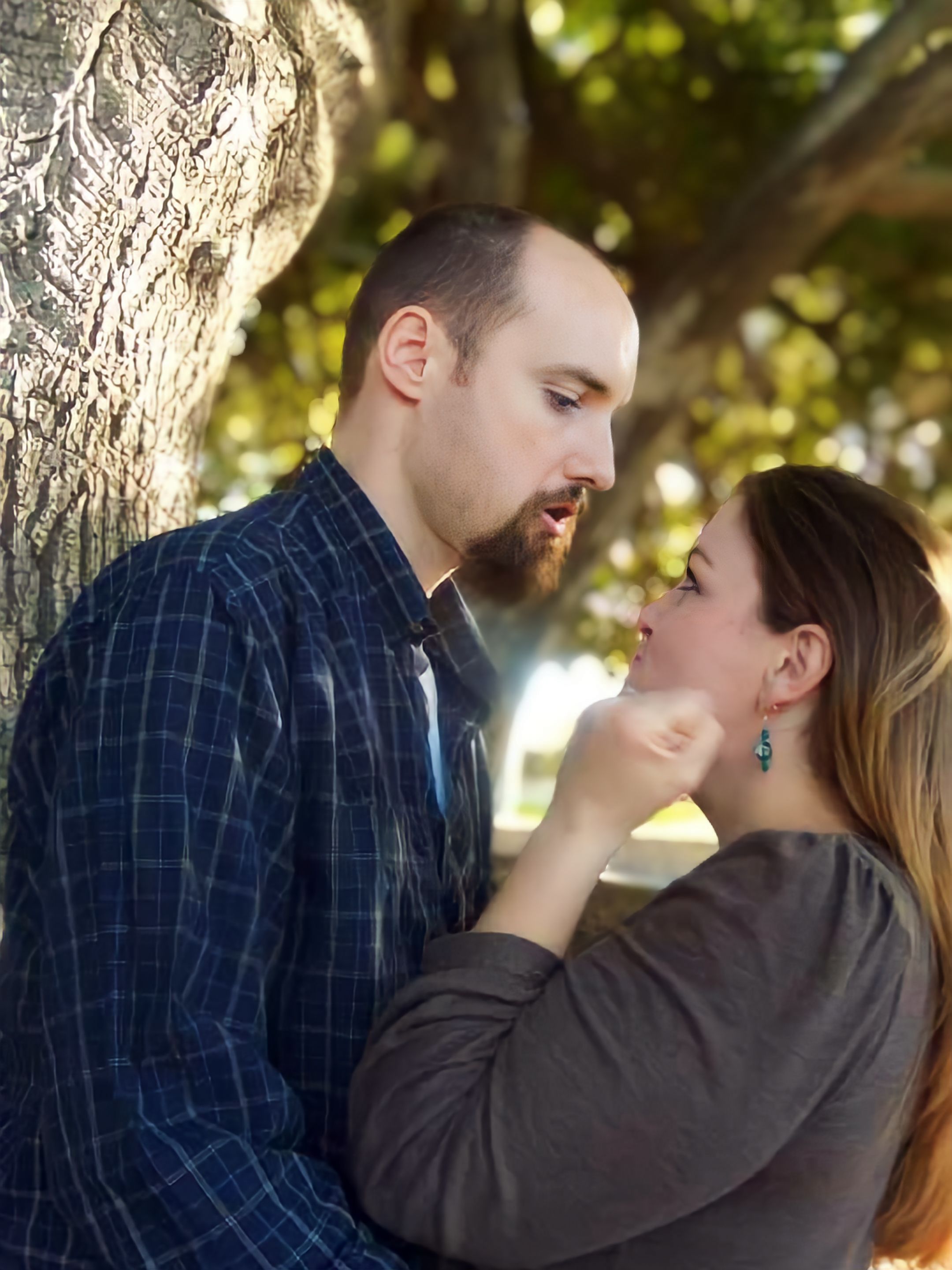 A romantic couple shares an intimate moment near a tree trunk on a sunny day, with soft bokeh lighting in the background.