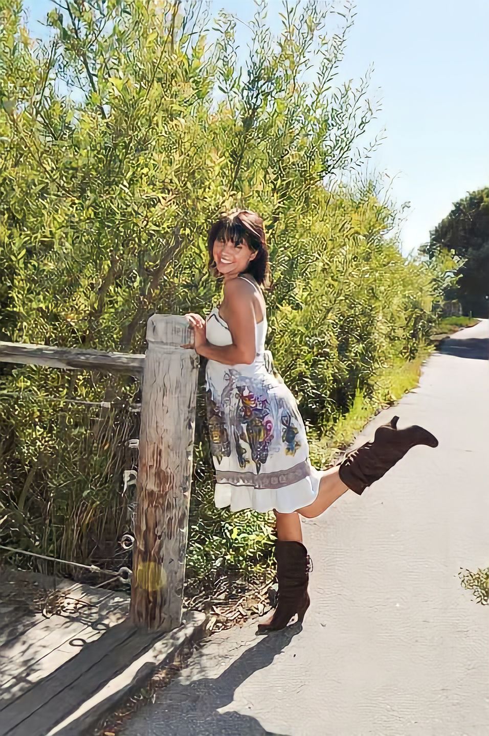 A sunlit path bordered by greenery showcases a whimsical fashion pose featuring a flowing dress and brown boots.