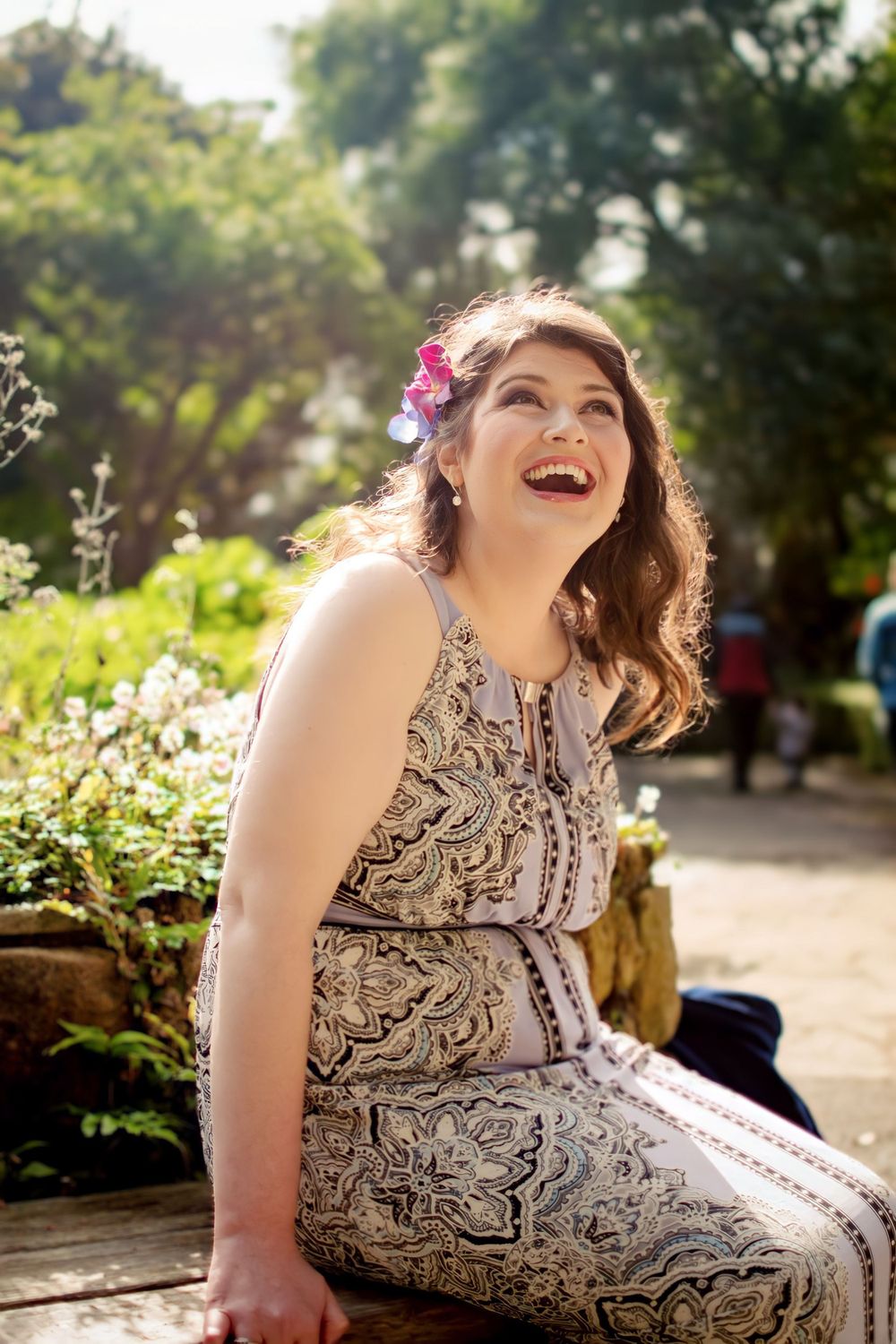 Joyful person in patterned dress and pink flower hair accessory laughs while sitting in sunny garden setting.