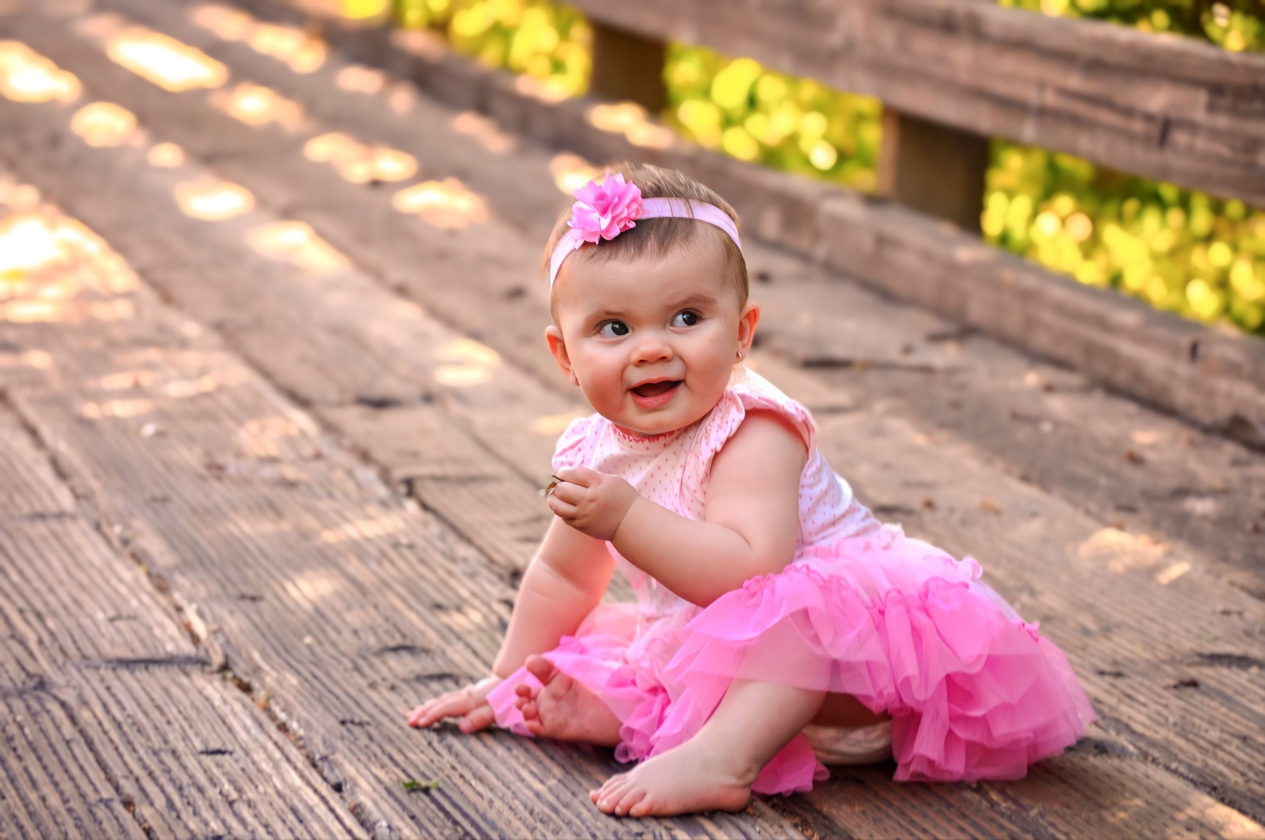 A smiling baby wearing a pink tutu and headband sits on wooden decking in warm outdoor lighting.