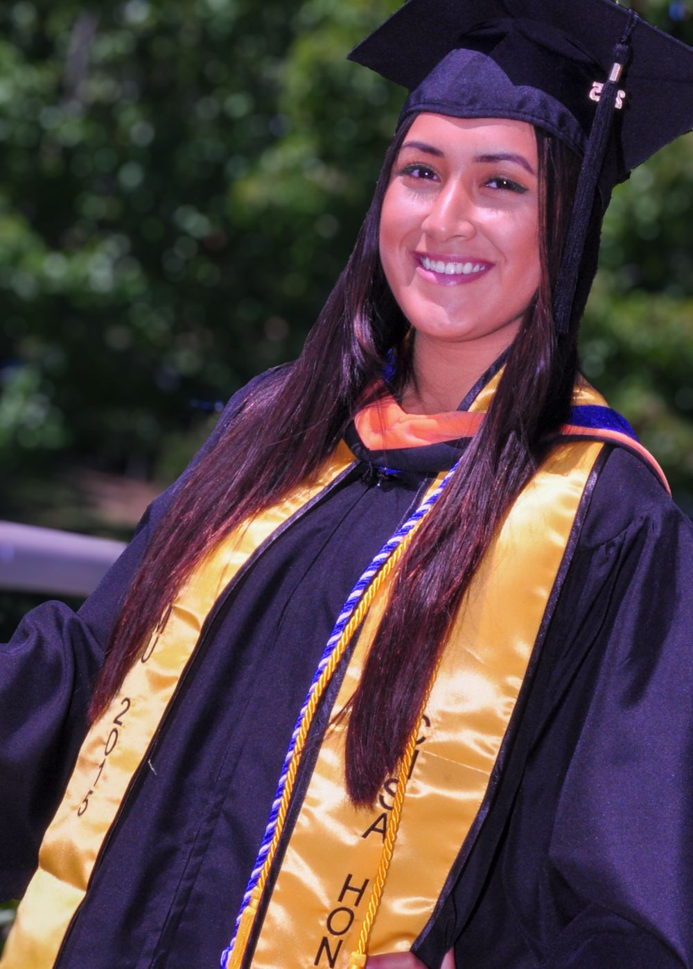 Graduate in black cap and gown with gold and orange honor stoles smiles brightly during commencement ceremony outdoors.