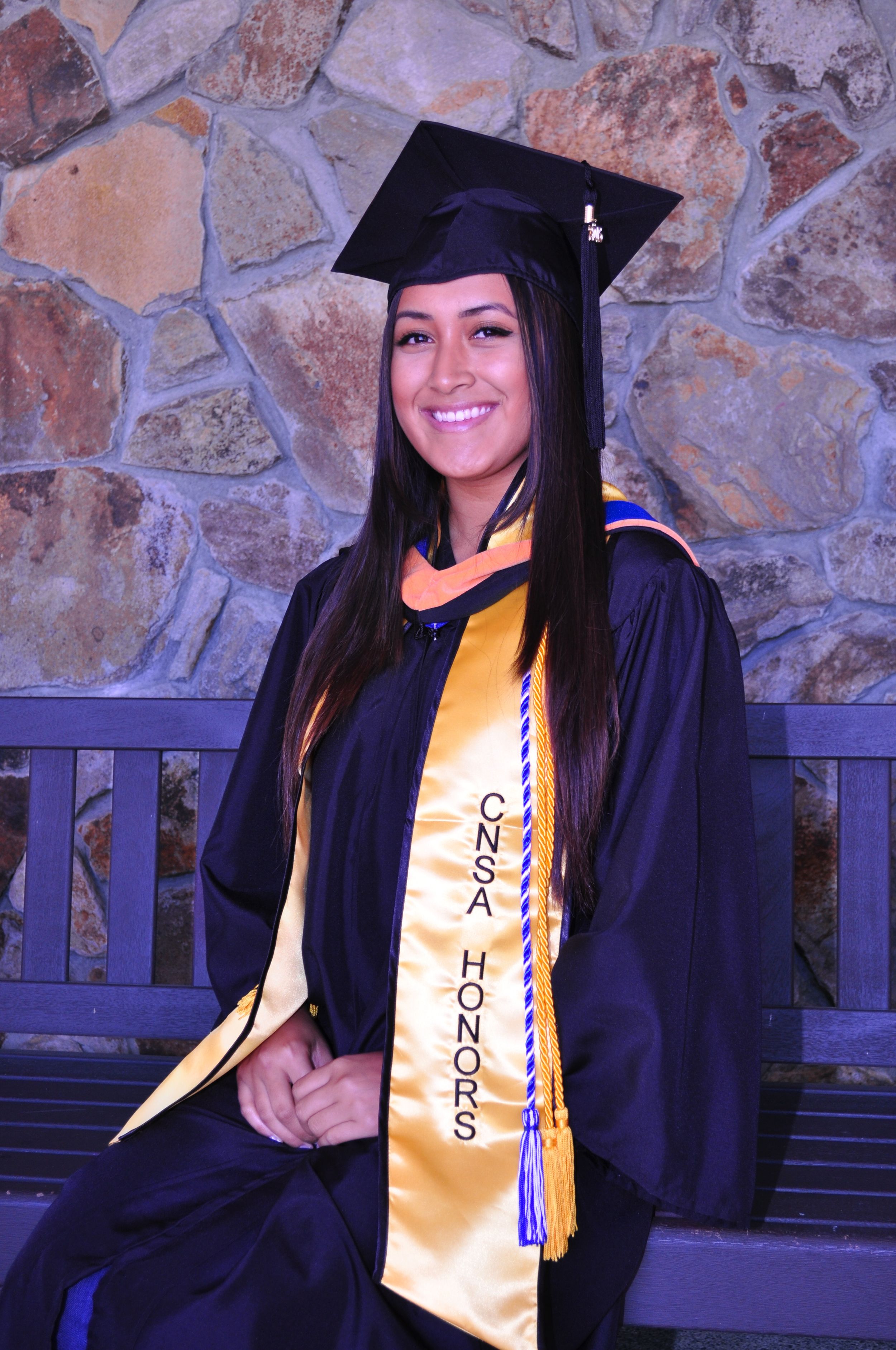 Graduate wearing traditional cap and gown with gold honors sash poses against stone wall background.