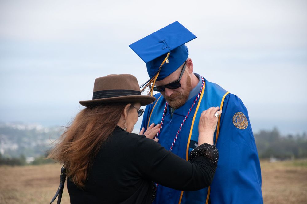 A student in blue graduation regalia and cap shares a tender moment with someone wearing brown hat outdoors.