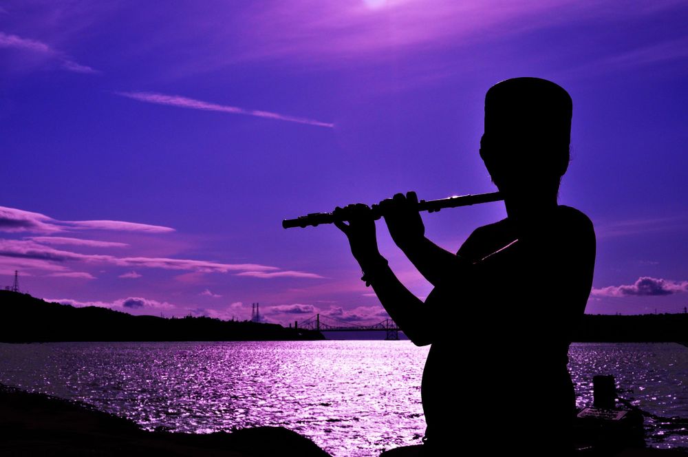 Silhouette of a person playing flute by the water during a vibrant purple sunset.