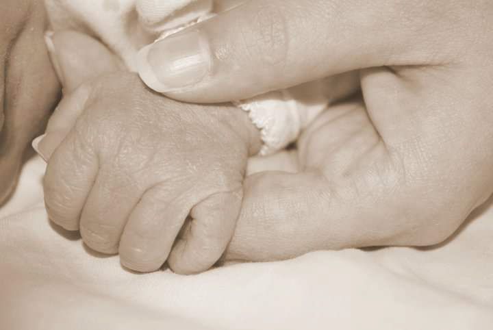 A gentle sepia-toned close-up photograph shows a tiny infant's hand grasping someone's finger.