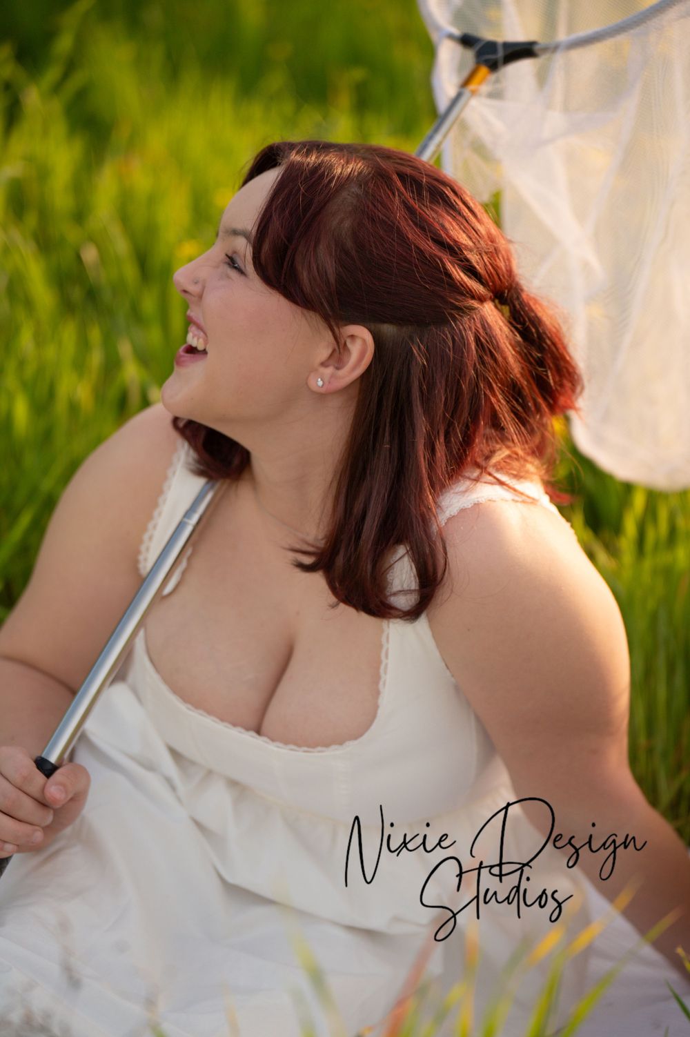 Woman in white dress laughs joyfully while sitting in sun-drenched grass during outdoor photo session.
