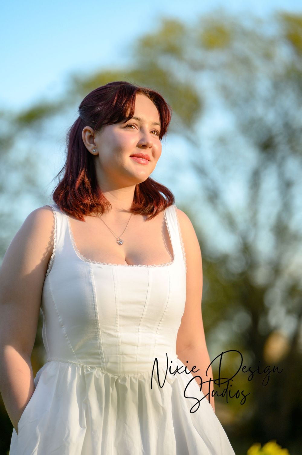 A person in a white sundress poses outdoors during golden hour with trees softly blurred in the background.