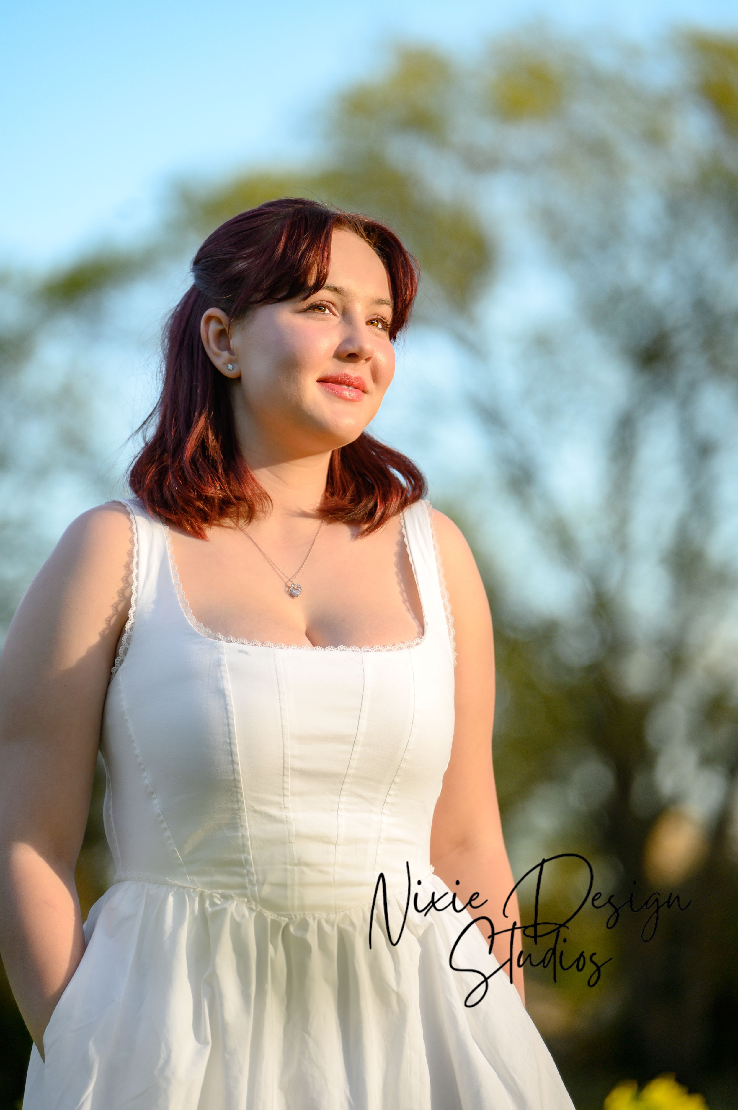 A person in a white sundress poses outdoors during golden hour with trees softly blurred in the background.