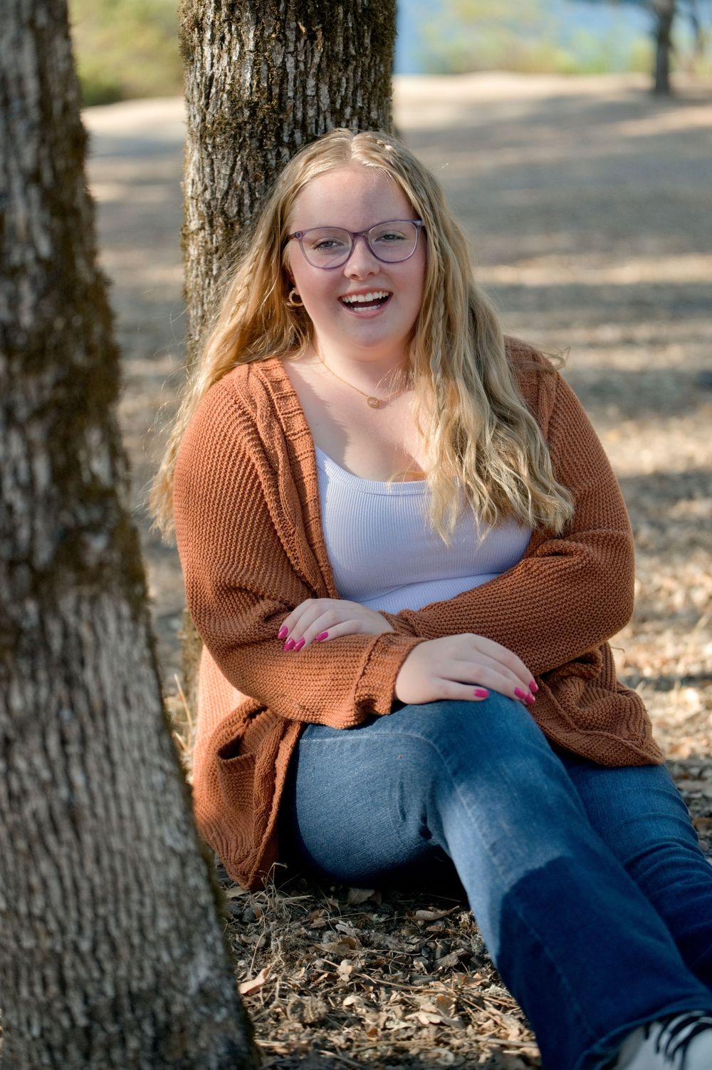 A person in a rust-colored cardigan and jeans sits smiling against a tree trunk on a sunny autumn day.