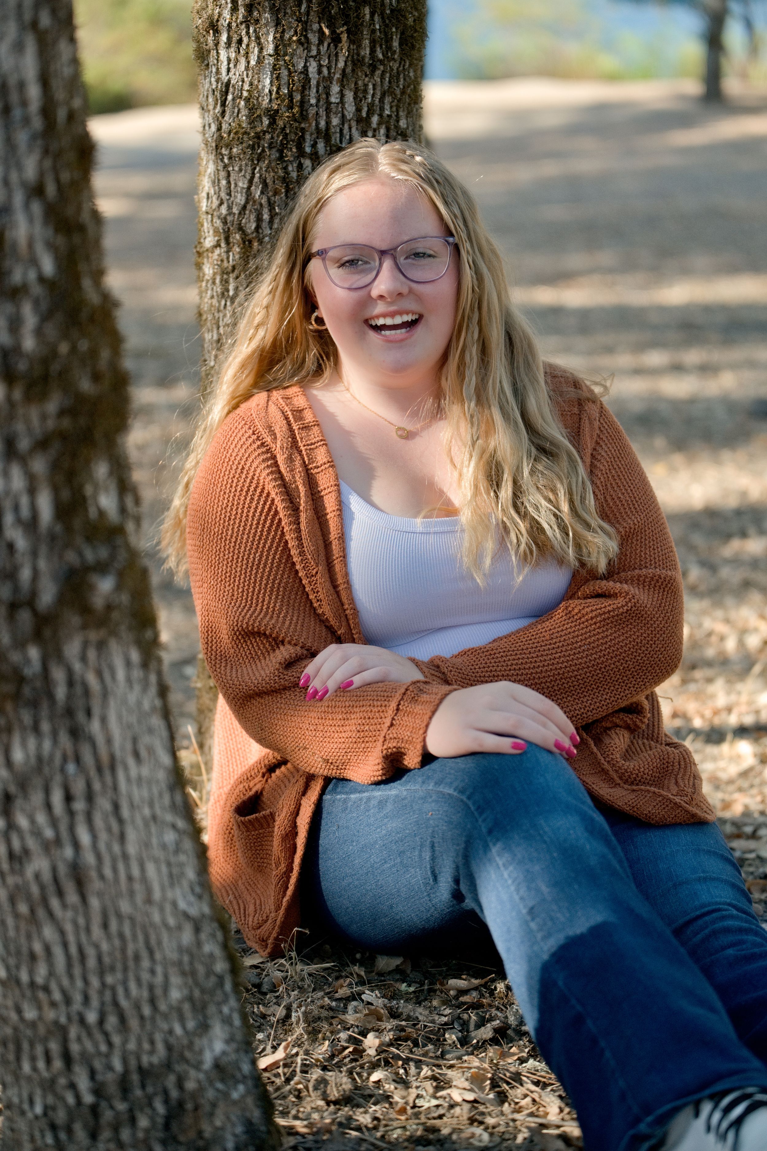 A person in a rust-colored cardigan and jeans sits smiling against a tree trunk on a sunny autumn day.