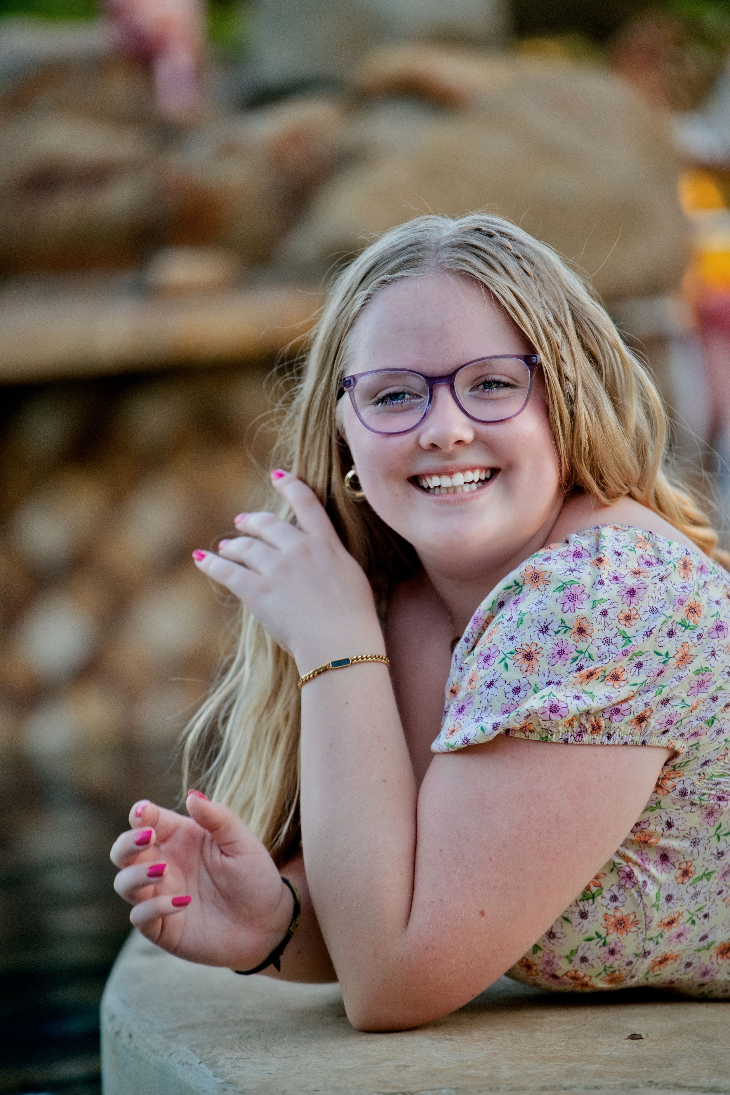 A joyful portrait of someone wearing glasses and a floral print top while leaning on a wood surface outdoors.
