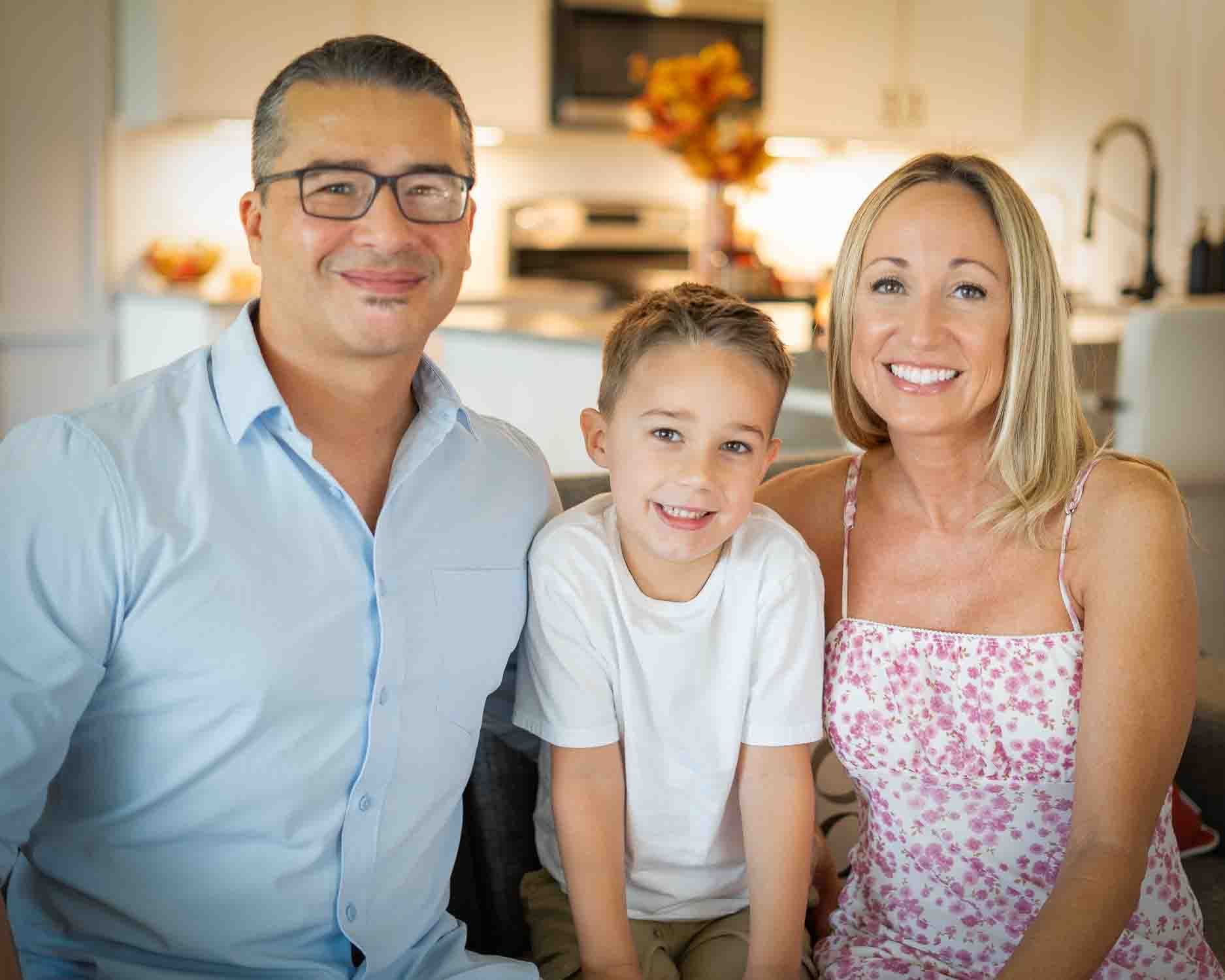 Family of three smiles together in bright kitchen with warm lighting.