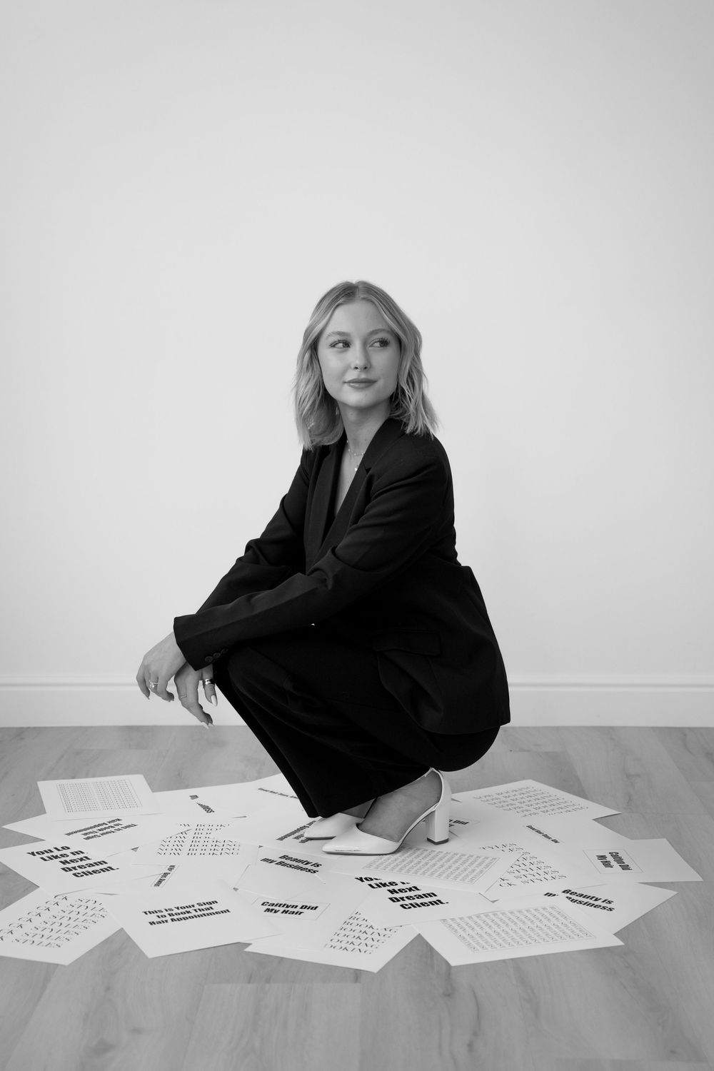 A figure in a black outfit sits on a white floor surrounded by scattered papers in a minimalist setting.