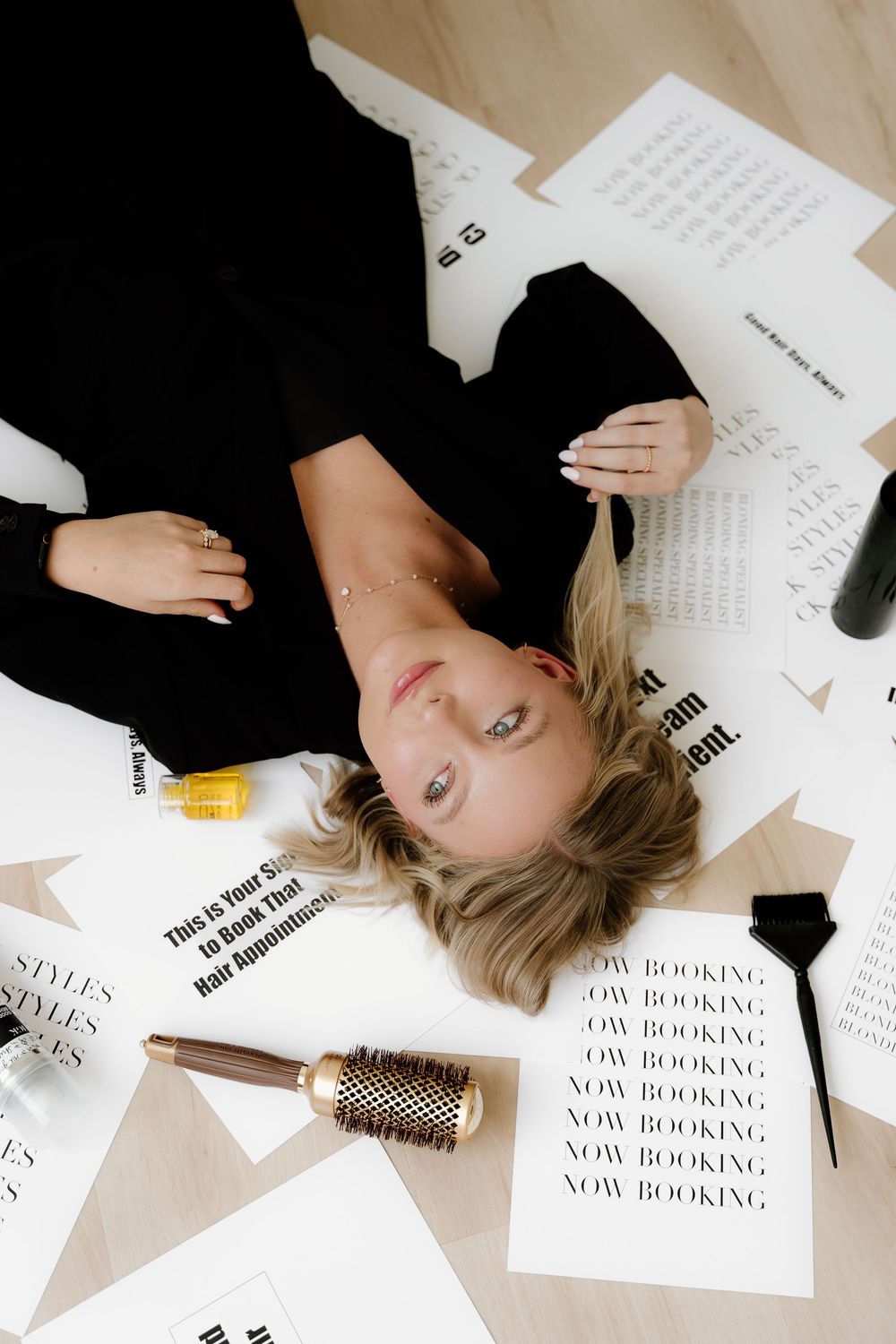 Person lying on floor surrounded by scattered papers with 'NOW BOOKING' text and a round hairbrush.